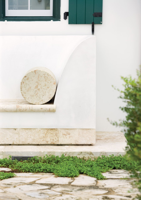 White stucco exterior wall with a curved built-in bench topped with travertine and a cylindrical stone accent. Green shutters flank a window above.