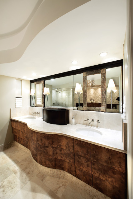 Modern bathroom with a curved vanity featuring a burl wood finish, marble countertop, and dual sinks. A large mirrored wall reflects a walk-in shower and decorative lamps.