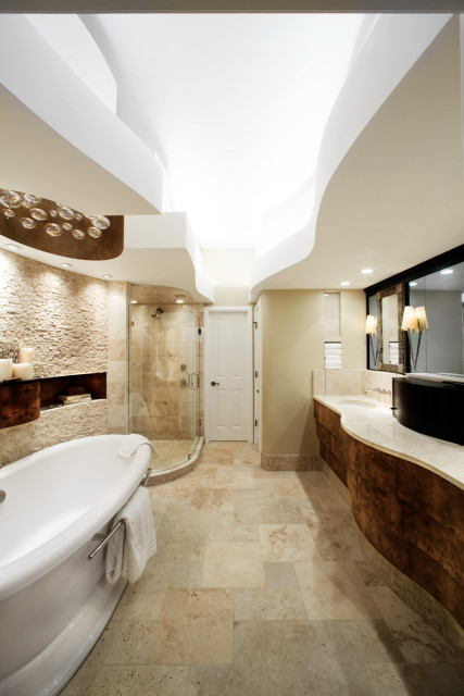 Luxury bathroom with freestanding white tub, stone tile walls, glass-enclosed shower, and curved wood vanity with marble countertop.