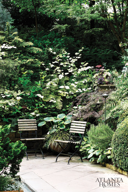 Stone patio with two wooden slatted chairs surrounded by lush green foliage, ferns, and flowering plants.