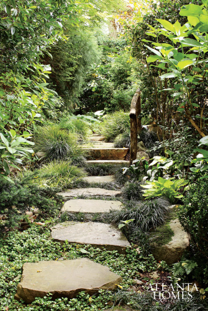 Stone stepping path winding through a lush garden with dense green foliage and ornamental grasses. A rustic wooden railing lines the right side of the path.