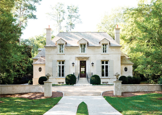 French country style stucco home with dormer windows, gray shutters, and a dark wood front door, surrounded by lush green trees and a manicured lawn.