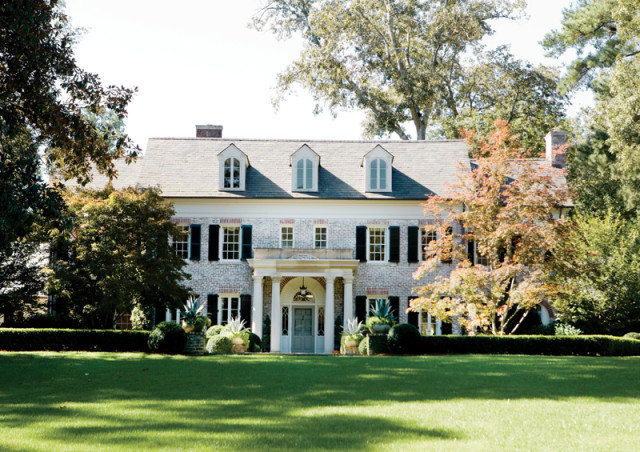 Federal-style white brick house with black shutters, dormer windows, and a columned portico. Lush green lawn and mature trees surround the property.