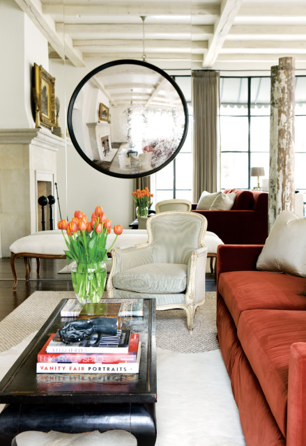 Living room with a large round mirror reflecting a fireplace, orange tulips on a coffee table, and rust-colored sofas.