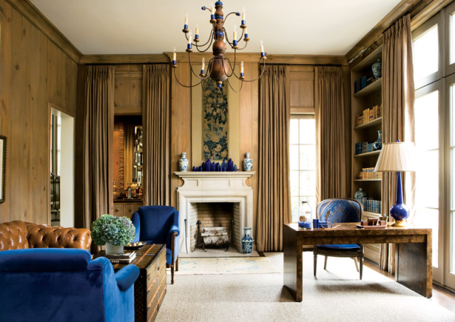Study with wood-paneled walls, a white fireplace, blue velvet armchairs, a leather sofa, a trunk coffee table, and a desk with a blue lamp.
