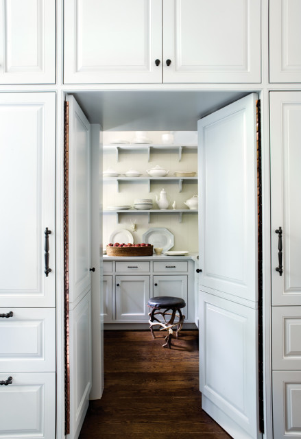 Pantry with white cabinetry and shelves stocked with white dishes and servingware. A woven basket of red apples sits on the counter, with a decorative stool nearby.