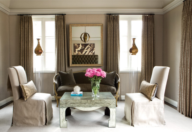 Living room with taupe walls, featuring a dark brown tufted sofa, two cream slip-covered chairs, a mirrored coffee table, and floor-to-ceiling patterned curtains.