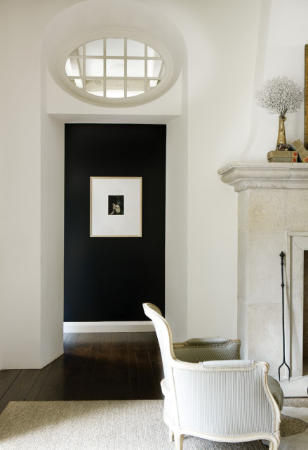 Hallway with black accent wall featuring framed art, leading to dark wood floors. A limestone fireplace and striped armchair are visible in the foreground.