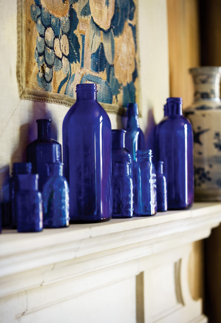 Collection of antique cobalt blue glass bottles of various sizes displayed on a white mantelpiece, with a tapestry in the background.