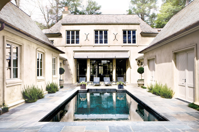 French country style home with stucco exterior, slate roof, and courtyard pool. Features include large windows, awnings, and manicured landscaping.