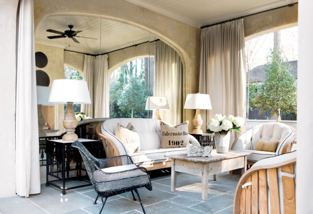 Sunroom with light-colored sofa and chairs, wooden coffee table, stone tile floor, and large arched windows with cream curtains.