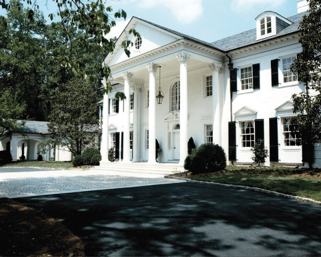 White neoclassical mansion with a grand portico supported by Corinthian columns, black shutters, and a slate roof, surrounded by mature trees and manicured landscaping.