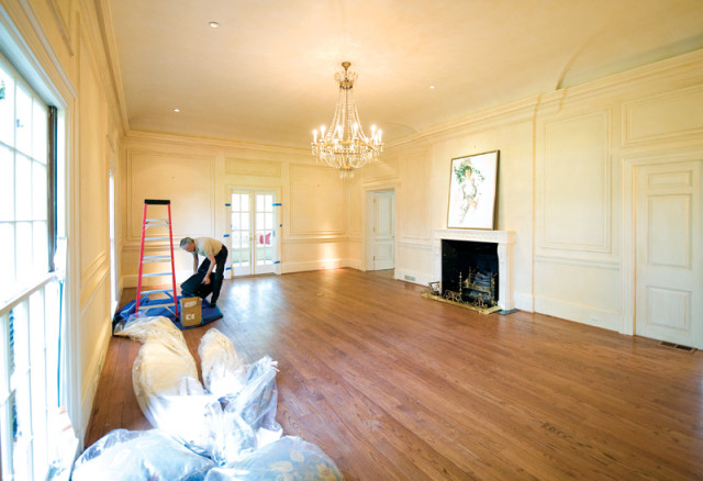Man unpacking in a large, empty living room with hardwood floors, a fireplace, a crystal chandelier, and French doors.