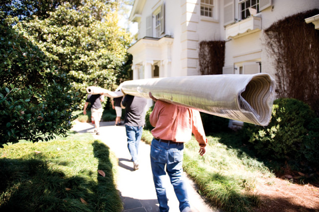 Three men carry a rolled carpet on their shoulders along a stone path next to a white house with green landscaping.