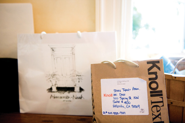 Two shopping bags on a wooden surface. The white bag on the left has a sketch of a doorway and plants. The brown bag on the right has the KnollText logo and a shipping label with an address and phone number.