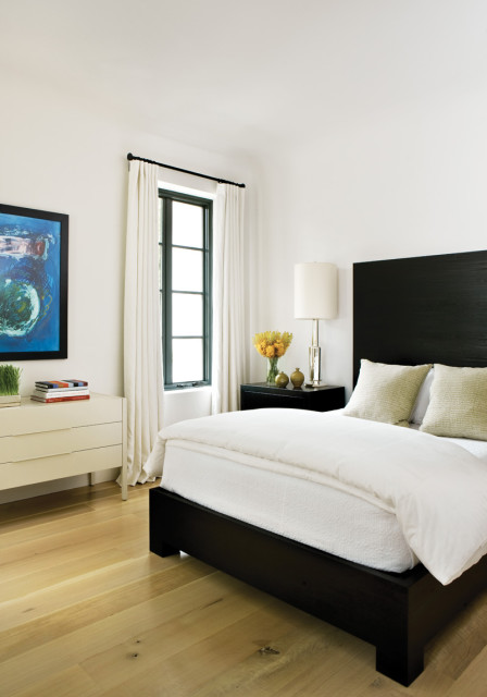 Bedroom with a dark wood platform bed, white bedding, two textured pillows, a black nightstand with a lamp and vase of yellow flowers, and a white dresser with a framed abstract painting.