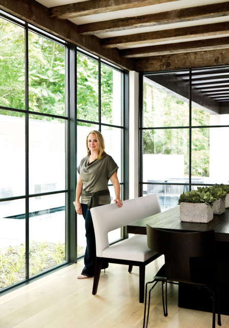 Woman standing by a large window in a dining room with a dark wood table, white banquette seating, and concrete planters with succulents.