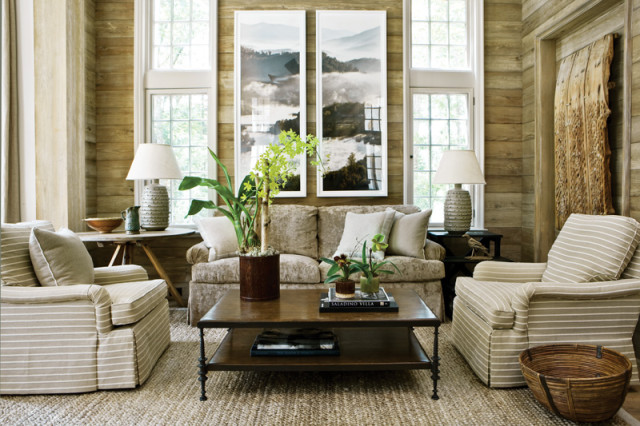 Living room with wood-paneled walls, two striped armchairs, a neutral sofa, a dark wood coffee table, and two framed landscape artworks above the sofa.