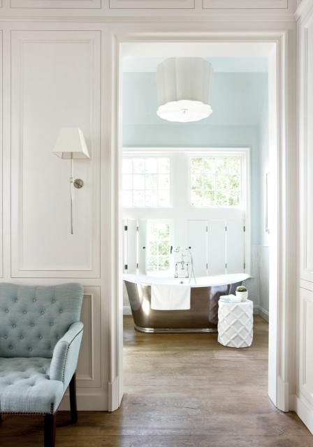 View through an archway into a light blue bathroom with a freestanding metallic tub, white shutters, and a large decorative ceiling light.