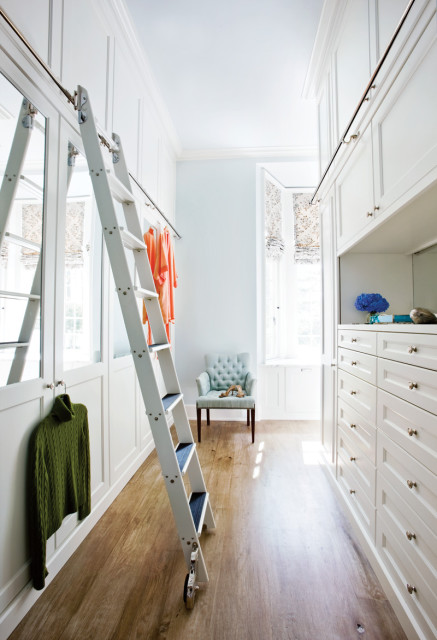 White walk-in closet with floor-to-ceiling custom cabinetry, a rolling ladder, tufted armchair, and hardwood floors.