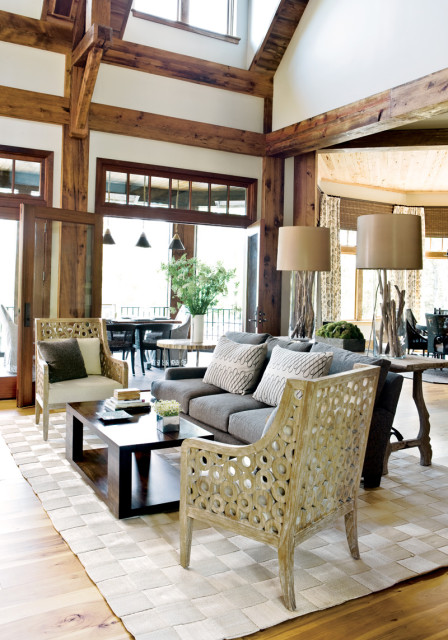 Living room with exposed wooden beams, gray sofa with patterned pillows, and two carved wooden accent chairs. A dark wood coffee table sits on a woven rug.