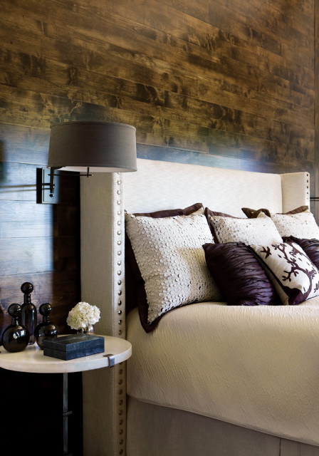 Bedroom with a king-sized bed featuring a tufted headboard, textured cream bedding, and an assortment of decorative pillows. A dark wood plank wall serves as a backdrop, complemented by a bedside table with decorative objects and a wall-mounted lamp.