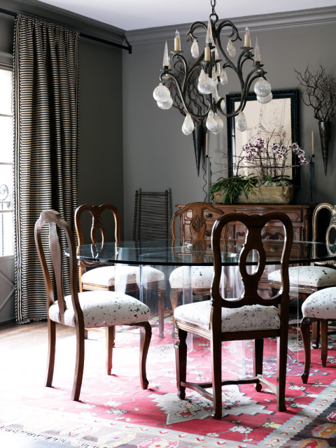 Dining room with dark gray walls, a glass-top dining table with wooden chairs upholstered in speckled fabric, a large ornate chandelier, and a patterned rug.