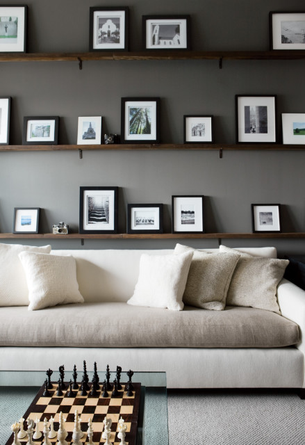Living room with a gray wall featuring three wooden shelves displaying framed black and white photographs and a white sofa with textured pillows. A chessboard sits on a glass coffee table in the foreground.