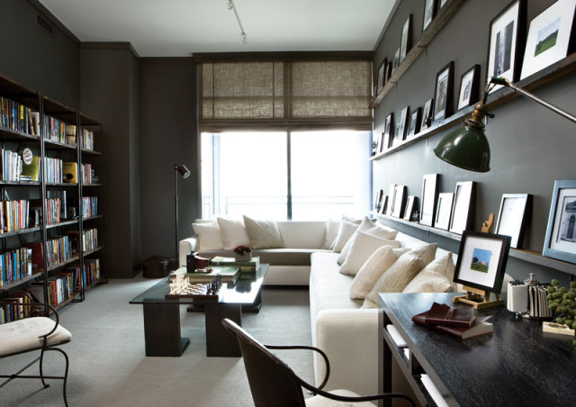 Living room with dark gray walls, floor-to-ceiling bookshelves, a white sectional sofa, glass coffee table with a chessboard, and a desk with framed art.