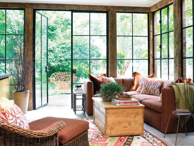 Sunroom with large windows overlooking a garden, featuring a rust-colored sectional sofa, a wooden chest coffee table, a wicker chair, and a colorful patterned rug.