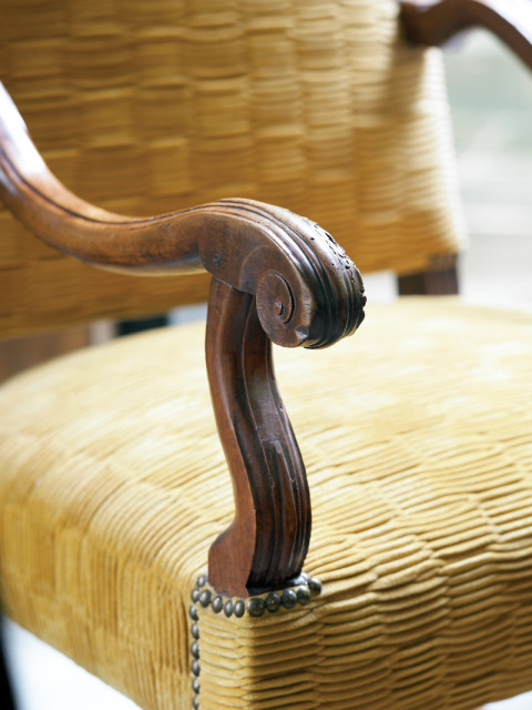 Close-up of a carved wooden armrest on a chair with yellow textured upholstery and decorative nailhead trim.