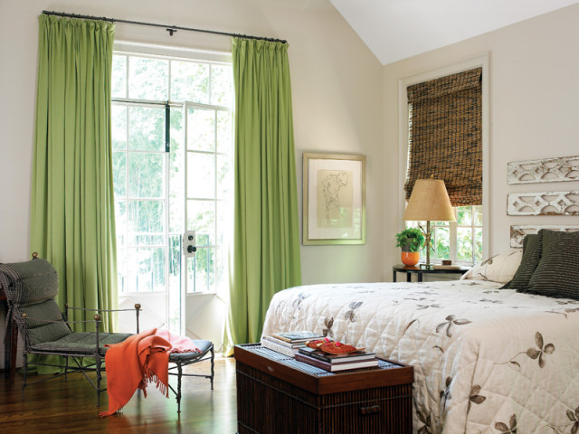 Bedroom with light green curtains, a floral comforter, a dark wood trunk, and a metal chair with an orange throw blanket.