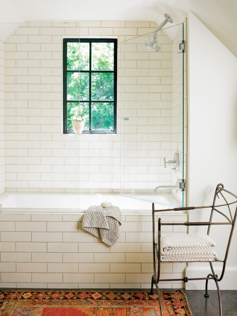 White subway tile bathroom with a black-framed window above a bathtub, glass shower enclosure, and a decorative metal chair holding towels.