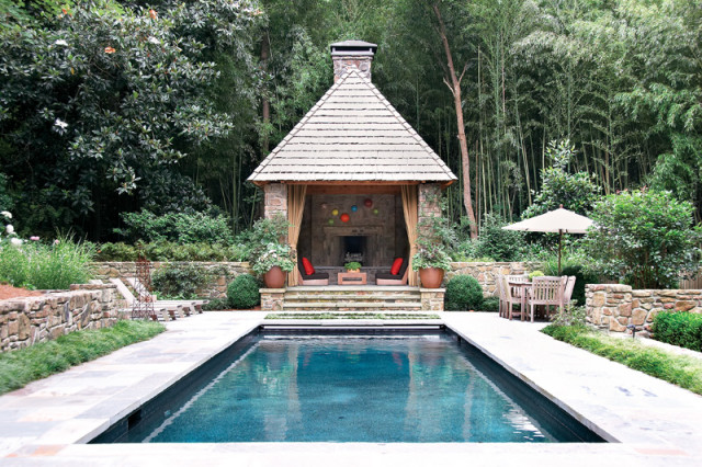 Rectangular swimming pool with stone patio leading to a covered outdoor living area with fireplace and seating, surrounded by lush bamboo and stone walls.