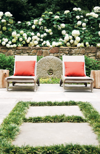 Two lounge chairs with orange cushions sit on a stone patio, backed by a stone wall and lush white hydrangeas. A large, circular stone feature is visible behind the chairs.