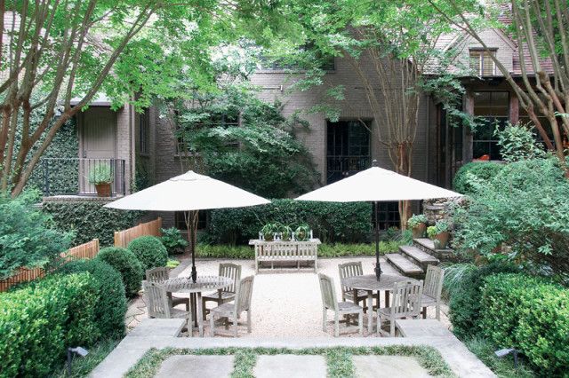 Courtyard with two round wooden tables and chairs under white umbrellas, surrounded by manicured boxwood hedges and gravel pathways. A brick building with dark windows is visible in the background.