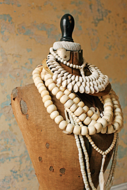 Close-up of a vintage mannequin torso covered in multiple strands of cream-colored beads and shell necklaces against a textured, distressed wall.