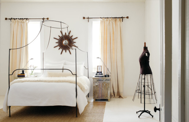 Bedroom with a black metal canopy bed, white quilted bedding, and a faux fur throw. A sunburst mirror hangs above the bed, and cream curtains frame the windows.