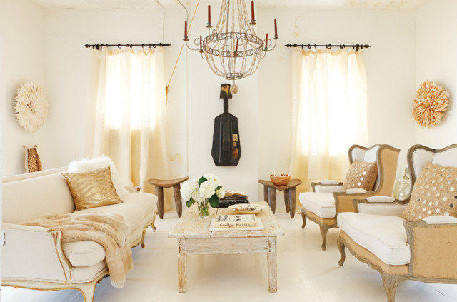 Living room with cream-colored furniture, a distressed wood coffee table, and two armchairs with mirrored accent pillows. Walls are white with sheer curtains and African-inspired wall decor.