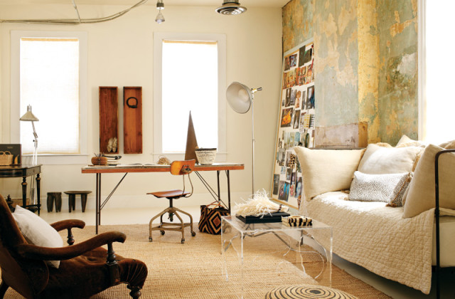 Eclectic studio space with a wooden desk, vintage chair, daybed with neutral pillows, and a wall collage of photos. Dominant colors are cream, brown, and muted green.