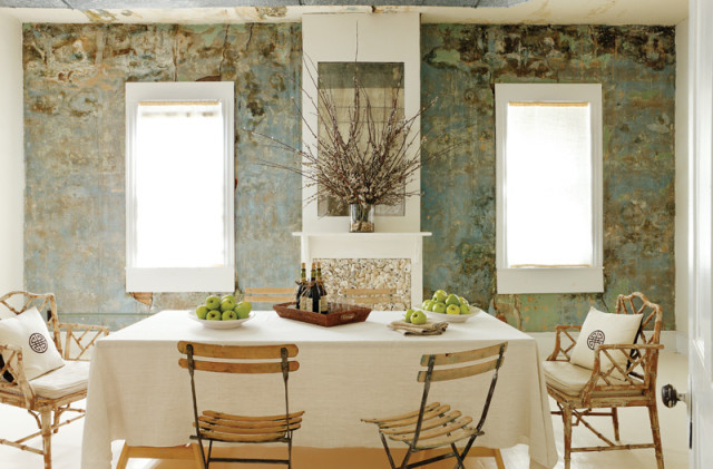 Dining room with distressed teal and brown walls, a white tablecloth-covered table set with green apples and wine bottles, and bamboo chairs with decorative pillows.