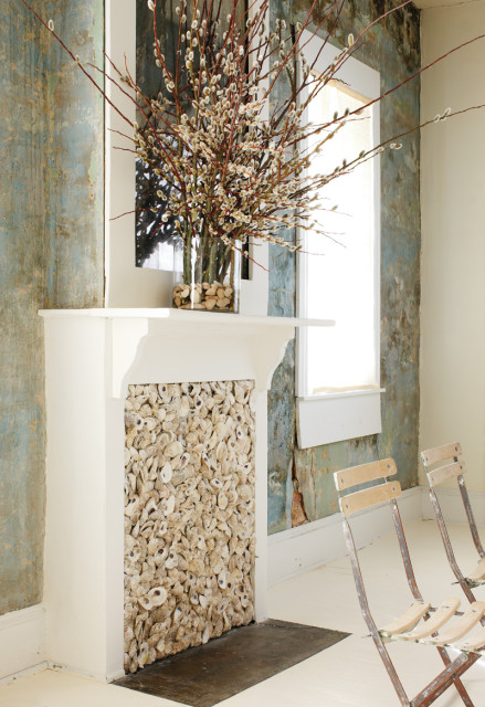 White fireplace with oyster shell surround, topped with a glass vase of pussy willow branches and stones. Walls have distressed blue and green finish.