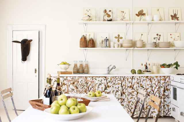 Cream kitchen with white cabinets, floral patterned skirted base cabinets, open shelving displaying artwork and dishware, and a wooden bull head mounted on a white door.