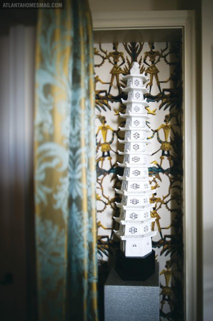 White pagoda sculpture displayed against patterned wallpaper featuring figures in gold and teal. A patterned curtain hangs to the left.