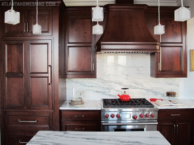 Dark cherry wood kitchen cabinets with marble countertops and backsplash, stainless steel range, and glass pendant lights.
