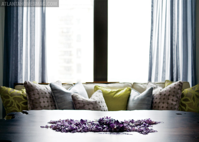 Dark wood table with amethyst crystal cluster centerpiece in front of a window with blue curtains and a sofa with patterned pillows.