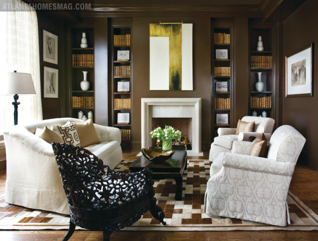 Brown living room with white tufted sofa, ornate dark wood chair, patterned armchairs, fireplace, and built-in bookshelves filled with books.