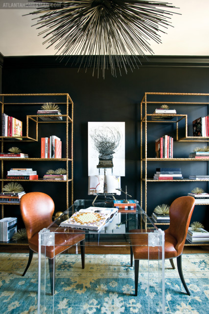 Black office with a clear acrylic desk, two brown leather chairs, gold shelving units filled with books, and a spiky black chandelier.