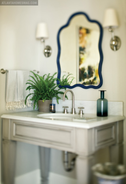 Powder room with a gray vanity, marble countertop, white sink, brushed nickel faucet, and a scalloped navy mirror. A potted fern and a dark green glass vase sit on the counter.