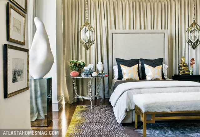 Bedroom with a large upholstered headboard, dark pillows with gold patterned accents, and a white fur bench. Neutral curtains hang behind the bed, and a geometric rug covers the floor.